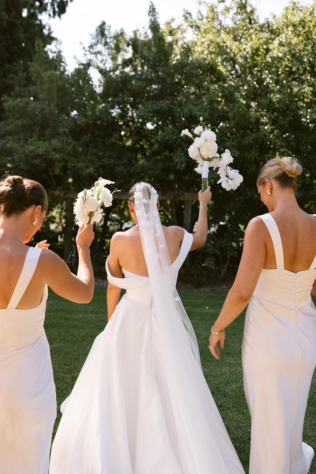A bride and two bridesmaids from behind, bouquets raised in the air