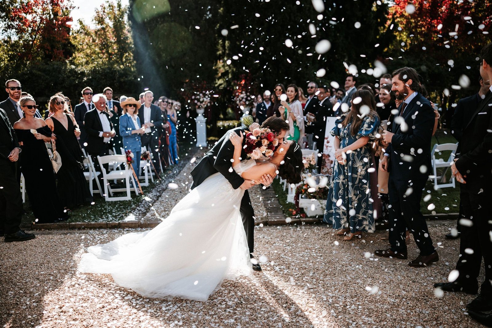 The groom dips the bride for a kiss as guests throw confetti along the gravel aisle