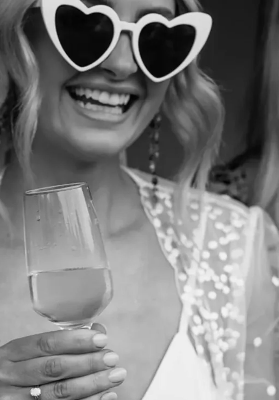 A bride laughing in heart-shaped sunglasses with a glass of champagne, in black and white