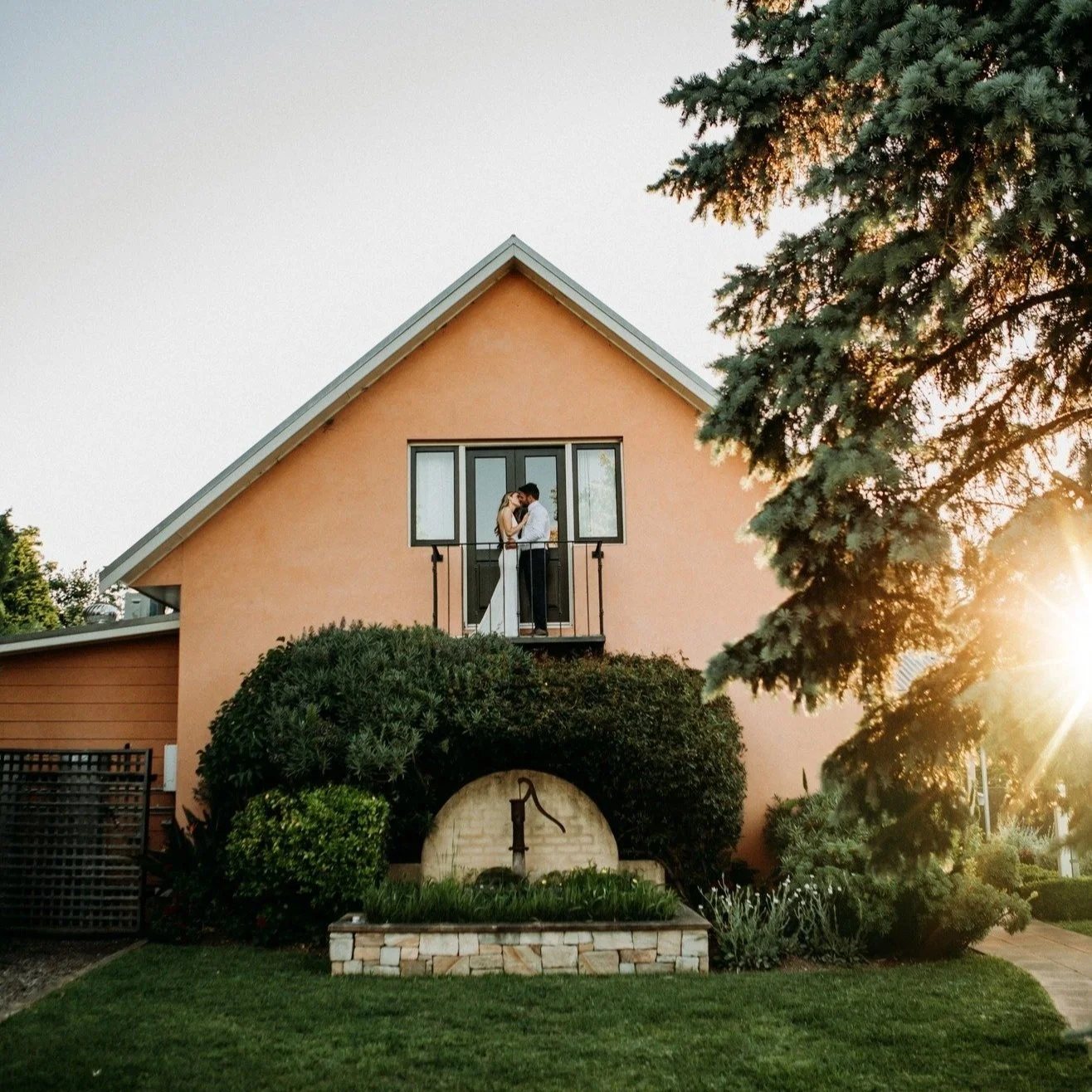 A couple kissing in the upstairs window of the terracotta cottage, sun flaring through the pines