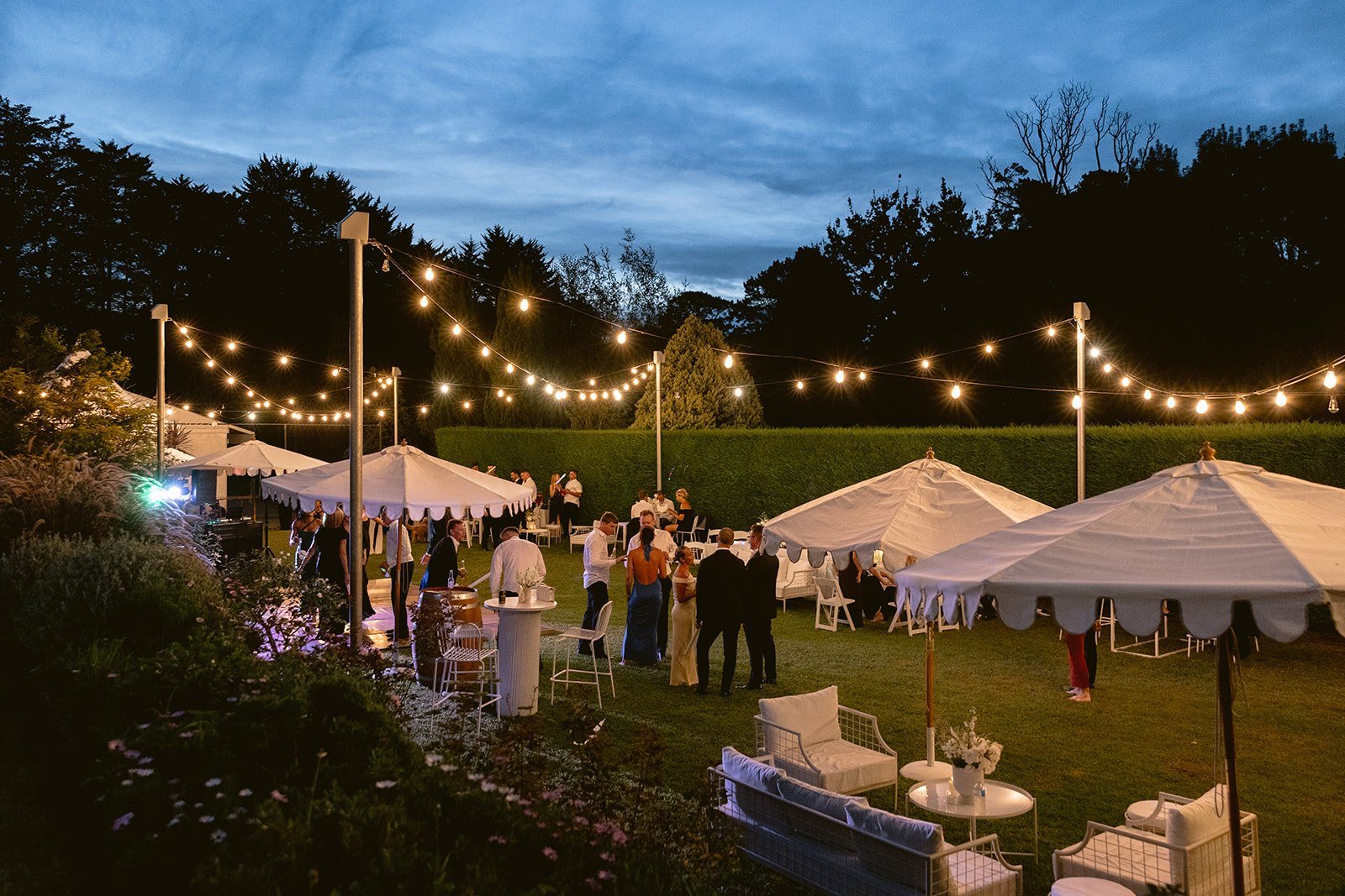 Guests on the lawn at dusk under festoon lights and white market umbrellas