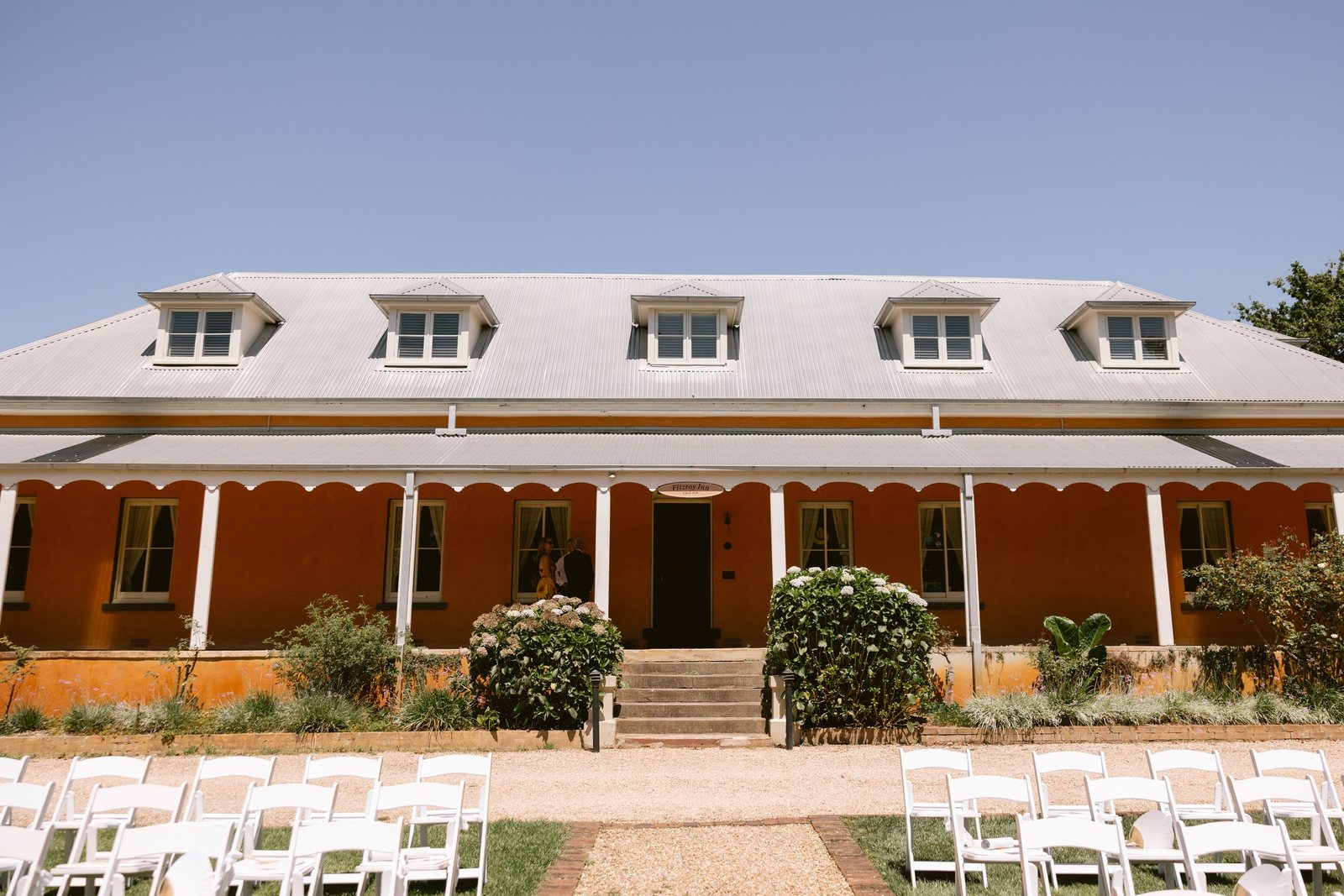 White ceremony chairs set on the lawn in front of the inn&rsquo;s long sandstone verandah