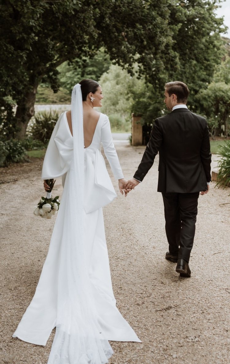 A couple walking hand in hand down the gravel drive under the trees, her long veil trailing